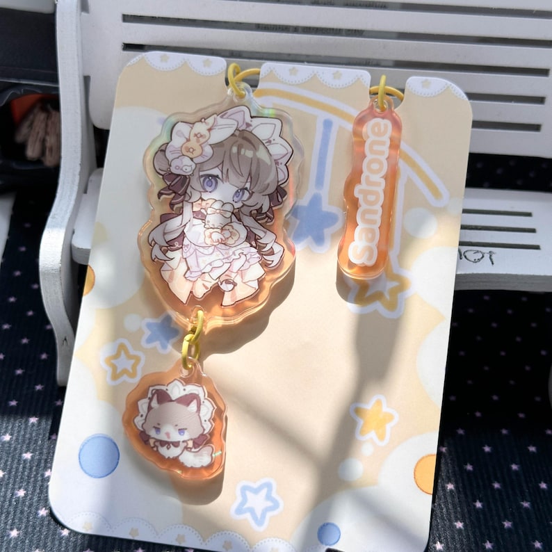 May include: An acrylic charm set featuring a character with brown hair in a frilled headpiece, a smaller cat-like creature charm, and a vertical orange tag with the text Sandrone, all attached to a light yellow backing card with star patterns.