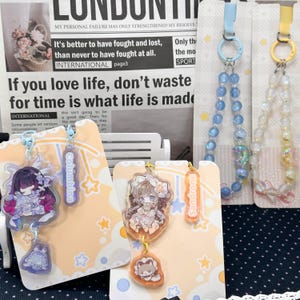 May include: Two acrylic charms featuring anime-style characters named Columbina and Sandrone, each attached to a backing card with star patterns. The Columbina charm is purple and blue, while the Sandrone charm is orange and brown, both with small animal pendants.