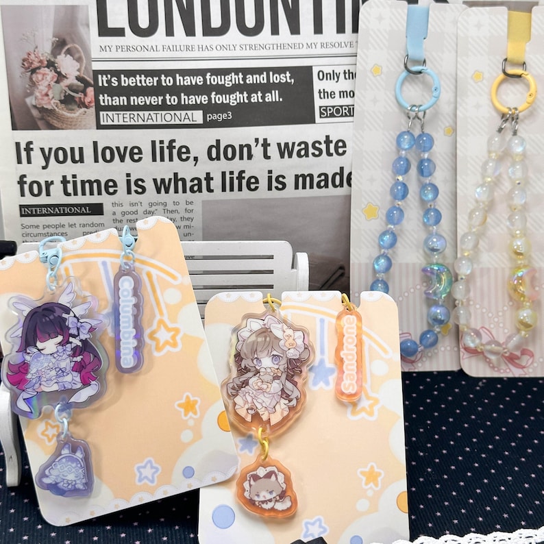 May include: Two acrylic charms featuring anime-style characters named Columbina and Sandrone, each attached to a backing card with star patterns. The Columbina charm is purple and blue, while the Sandrone charm is orange and brown, both with small animal pendants.