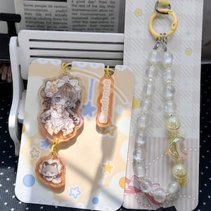 May include: An acrylic charm set featuring an anime-style character in a frilled outfit, a smaller charm with the text Sandrone, and a matching charm with a cat-like figure. A separate beaded phone strap features iridescent beads and a crescent moon charm.