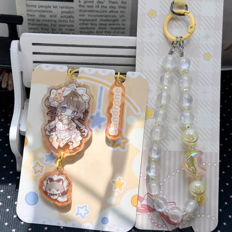 May include: An acrylic charm set featuring an anime-style character in a frilled outfit, a smaller charm with the text Sandrone, and a matching charm with a cat-like figure. A separate beaded phone strap features iridescent beads and a crescent moon charm.