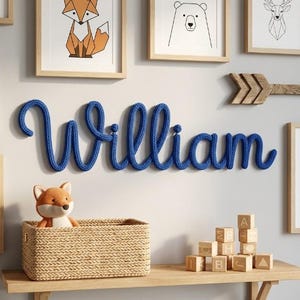 Pode incluir: Um letreiro de parede feito de corda tricotada azul que soletra o nome William em letra cursiva. O letreiro está montado numa parede rodeado por ilustrações de animais emolduradas, uma seta de madeira, um cesto entrançado com uma raposa de peluche e blocos de madeira.