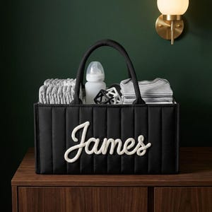 Puede incluir: Una cesta de almacenamiento rectangular acolchada de color negro con dos asas, que presenta el nombre James en letras blancas estilo cuerda en la parte frontal. La cesta contiene ropa plegada a rayas grises y blancas, un biberón y un juguete negro.