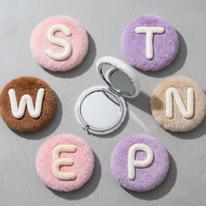 Pode incluir: Um conjunto de seis espelhos de bolso redondos de pelo sintético felpudo em várias cores pastel e neutras, cada um com uma letra branca grande costurada na frente. As letras são S, T, W, N, E e P. Um espelho está aberto, revelando a superfície refletora.