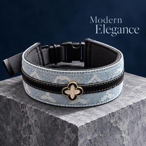 Puede incluir: Un collar de mezclilla azul claro con un patrón blanco repetitivo, una franja horizontal negra central y un emblema floral dorado de cuatro pétalos con un centro blanco. El collar incluye una hebilla de plástico negro y el texto Modern Elegance.
