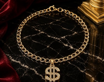 Puede incluir: Un collar de cadena en tono dorado con un colgante de signo de dólar incrustado con pequeñas piedras transparentes. El collar está construido con eslabones ovalados entrelazados e incluye un cierre de mosquetón en la parte posterior.