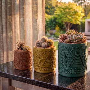 以下が含まれることがあります： テラコッタ、イエロー、ダークグリーンの3つの円筒形プランター。それぞれに階段状のピラミッドとドラゴンのレリーフが施されています。プランターには様々な多肉植物やサボテンが植えられており、左から右へサイズが大きくなっています。