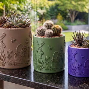 May include: Three cylindrical planters in beige, sage green, and purple, each featuring embossed designs of a windmill, a woven basket, wheat stalks, flowers, and bees. The planters hold various succulents and cacti with small pebbles on top.