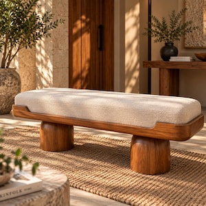 Puede incluir: Un banco rectangular con un cojín grueso de tela texturizada color crema, encajado en un marco de madera de tono oscuro. El asiento se apoya sobre dos patas de madera cilíndricas, redondeadas y robustas que ofrecen una base estable.