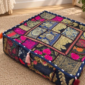 May include: A square floor cushion featuring a patchwork design of various fabrics in black, gold, and hot pink. The surface is decorated with intricate floral embroidery, metallic gold thread patterns, and small white tassels along the edges.