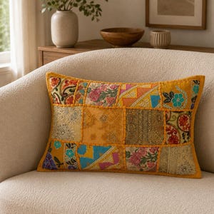 Può includere: Un cuscino rettangolare caratterizzato da un design patchwork realizzato con vari tessuti nei toni del giallo, arancione, blu e rosa. La superficie presenta intricati ricami floreali, accenti in filo metallico e cuciture decorative tra i pannelli.
