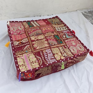 May include: A square floor cushion featuring a patchwork design of various red, gold, and pink fabrics with intricate embroidery, metallic thread detailing, and small mirrors. The edges are finished with a dark red border and colorful tassels.