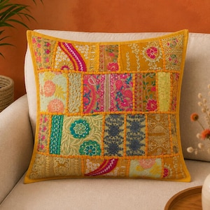 May include: A square throw pillow featuring a patchwork design of various embroidered fabrics in shades of mustard yellow, hot pink, teal, and gold. The surface is composed of rectangular panels with intricate floral and paisley patterns.