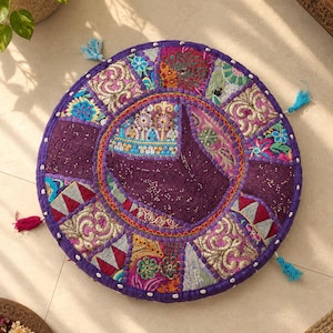 May include: A round floor cushion featuring a patchwork design with purple fabric as the base. The surface is decorated with intricate embroidery, sequins, and small white shells along the edge. Four small tassels in blue and pink are attached.