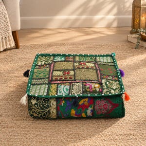 May include: A square floor cushion featuring a patchwork design of various green fabrics with gold embroidery, floral patterns, and metallic accents. The edges are finished with white pom-pom trim and small colorful tassels at each corner.