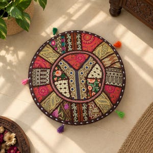 Könnte beinhalten: Ein rundes Bodenkissen mit einem Patchwork-Design aus bestickten Stoffbahnen in Rot-, Gold-, Grün- und Rosatönen. Das Kissen hat einen dunkelbraunen Rand mit weißen Nähten und kleinen bunten Quasten, die am äußeren Rand befestigt sind.