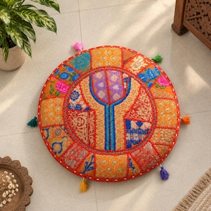 May include: A round floor cushion featuring a vibrant patchwork design with various floral and geometric patterns in shades of orange, red, blue, and purple. The cushion is accented with small colorful tassels along the outer edge.