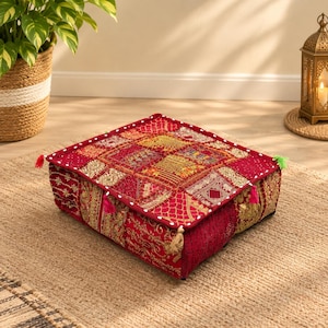Puede incluir: Un cojín de suelo cuadrado con un diseño de patchwork en tonos rojo intenso y dorado. La superficie está decorada con bordados intrincados, detalles en hilo metálico y pequeñas borlas en las esquinas. Los bordes tienen un ribete blanco.