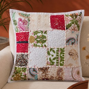 Op de afbeelding: Een vierkant sierkussen met een patchworkontwerp van diverse geborduurde stoffen vierkanten in tinten rood, groen, wit en goud. De vierkanten zijn verbonden door zichtbare witte stiksels, wat zorgt voor een textuurrijk oppervlak met bloemmotieven.