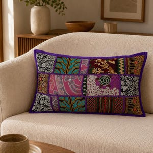 Pode incluir: Uma almofada retangular com um design de patchwork vibrante composto por vários tecidos bordados em tons de roxo, rosa, azul-petróleo e preto. A almofada é contornada por um rebordo de tecido roxo sólido e exibe padrões florais e geométricos intrincados.