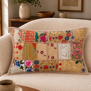 Puede incluir: Un cojín rectangular con un diseño de patchwork compuesto por varios cuadrados de tela. La superficie muestra una mezcla de bordados florales, abalorios, lentejuelas y encaje en colores vibrantes como rojo, rosa, azul y dorado sobre una base beige.