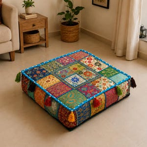 Op de afbeelding: Een vierkant vloerkussen met een patchworkontwerp van diverse kleurrijke stoffen met ingewikkelde bloemen- en paisleypatronen. De randen zijn afgewerkt met felblauwe biezen en geaccentueerd met kleine kwastjes in groen, bruin en rood.
