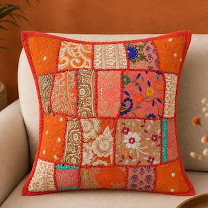 May include: A square throw pillow featuring a patchwork design of various fabrics in shades of orange, pink, and cream. The surface is decorated with intricate embroidery, floral patterns, and small reflective sequins, all framed by a red border.