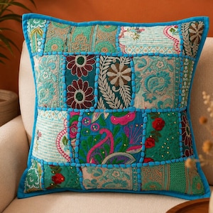Op de afbeelding: Een vierkant sierkussen met een patchworkontwerp van verschillende teal, groene en blauwe stoffen panelen. De panelen zijn versierd met ingewikkeld bloemen- en paisley-borduurwerk in rood, roze en wit, allemaal verbonden door felblauwe stiksels.