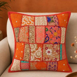 May include: A square throw pillow featuring a patchwork design of various fabric squares in shades of orange, pink, and gold. The fabric squares are decorated with intricate embroidery, floral patterns, and small metallic sequins.