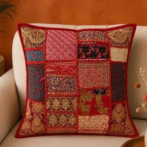 May include: A square throw pillow featuring a patchwork design of various red, gold, and dark-toned fabric panels. The surface is decorated with intricate embroidery, small reflective mirror accents, and metallic beadwork, finished with a red border.