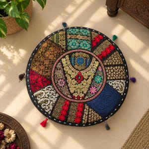 Puede incluir: Un cojín de suelo redondo con un diseño de patchwork de paneles de tela bordados en negro, rojo, azul, verde y dorado. El círculo central muestra un intrincado bordado de hilo dorado floral y geométrico, acentuado por pequeñas borlas coloridas.