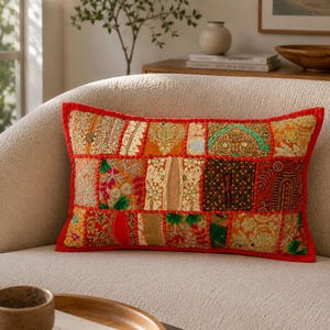 Könnte beinhalten: Ein rechteckiges Zierkissen mit einem Patchwork-Design aus verschiedenen bestickten Stoffen in Rot-, Orange-, Gold- und Grüntönen. Das Kissen ist mit einer leuchtend roten Stoffkante versehen und zeigt aufwendige Metallfadenstickereien und Pailletten.