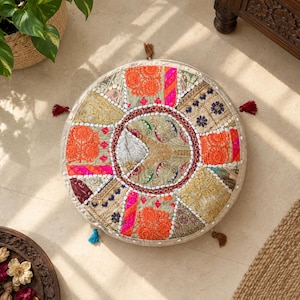 Puede incluir: Un cojín de suelo redondo con un diseño de patchwork de paneles de tela bordados en tonos vibrantes de naranja, rosa intenso, dorado y verde salvia. El cojín está decorado con pequeñas cuentas blancas, detalles de hilo metálico y borlas.