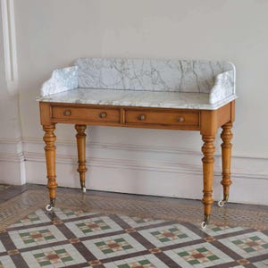 May include: A wooden console table featuring a white marble top with grey veining and a matching marble backsplash. The table has two drawers with small round knobs, four turned legs, and is supported by small metal casters at the base.