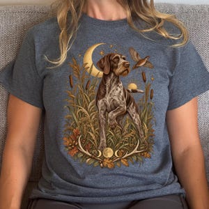 May include: A heathered dark gray t-shirt featuring a graphic illustration of a brown and white German Shorthaired Pointer standing in tall grass. The design includes a crescent moon, a flying bird, cattails, oak leaves, and antlers.