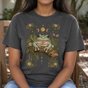 May include: A charcoal grey t-shirt featuring a central graphic of a green frog perched on a mossy branch. The design includes white water lilies, green leaves, mushrooms, a dragonfly, and golden celestial moon phases and stars surrounding the frog.