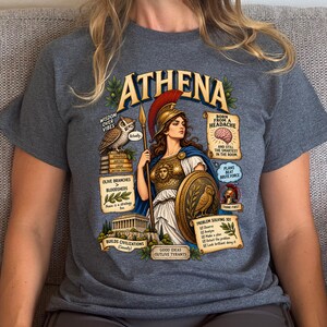 May include: A charcoal gray t-shirt featuring a graphic of Athena with a helmet, spear, and shield. The design includes an owl, a temple, and text boxes with phrases like "Wisdom over vibes," "Born from a headache," and "Problem solving 101."