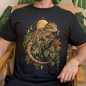 May include: A black short-sleeved t-shirt featuring a graphic print of a crested gecko perched on a branch. The design includes botanical elements, a golden moon, and celestial star symbols surrounding the lizard in shades of brown and green.