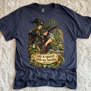 Op de afbeelding: Een donkergrijs t-shirt met een afbeelding van een persoon met een hoed met brede rand, versierd met bloemen en een halve maan, die een kleine vogel vasthoudt bij een bamboe waterornament. Onder de illustratie staat op een banier de tekst on a quest with my familiar.