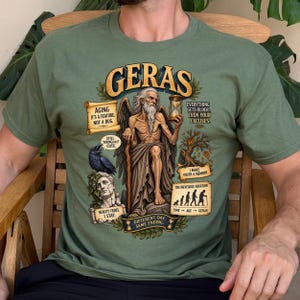 May include: An olive green t-shirt featuring a graphic of Geras, the personification of old age, with a long beard, wings, and an hourglass. The design includes text such as 'Aging it's a feature, not a bug', 'Still young? Cute', and 'The inevitable equation'.