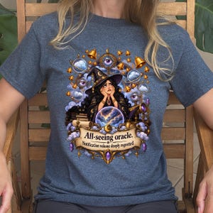 Może przedstawiać: Melirowany niebieski t-shirt z grafiką przedstawiającą czarownicę z długimi ciemnymi włosami i w spiczastym kapeluszu, otoczoną lewitującymi oczami, dzwonkami i chmurami. Pod czarownicą znajduje się kryształowa kula i baner z napisem All-seeing oracle. Notification volume deeply regretted.