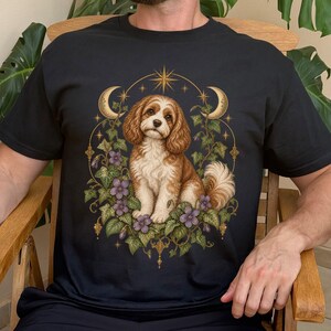 Può includere: Una t-shirt nera con la grafica di un cane spaniel marrone e bianco seduto tra foglie di edera verde e fiori viola. Il disegno è incorniciato da un bordo circolare color oro con due lune crescenti e una stella nella parte superiore.
