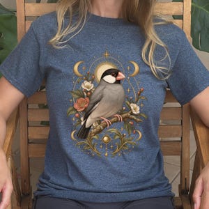 May include: A heather blue t-shirt featuring a graphic print of a Java sparrow perched on a branch. The bird is surrounded by a circular design with flowers, leaves, crescent moons, and a sunburst, all rendered in shades of gold, pink, and white.