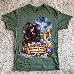 Op de afbeelding: Een olijfgroen t-shirt met een afbeelding van een heks in een paarse hoed en gewaad die een staf vasthoudt, vergezeld door een zwarte kat, servertorens en rollen. De tekst luidt: "Summoning servers also summons the invoice."