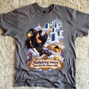 May include: A charcoal gray t-shirt featuring a graphic of a witch in a black dress and hat holding a glowing staff. She stands among clouds with floating towers and a golden pot of coins. The text reads: Multiplying towers faster than the treasury refills.