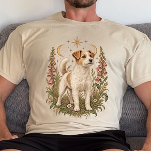 May include: A beige t-shirt featuring a graphic print of a white and brown Jack Russell Terrier standing in a patch of grass and flowers. The design includes two crescent moons, a central star, and tall pink foxglove flowers surrounding the dog.
