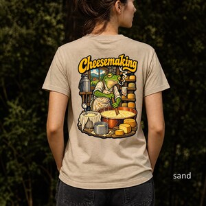 Op de afbeelding: Een crèmekleurig t-shirt met een afbeelding op de achterkant van een groene kikker in een schort die in een grote koperen pan roert. De tekst "Cheesemaking" staat erboven, met planken vol kaaswielen en een melkbus in de achtergrond.