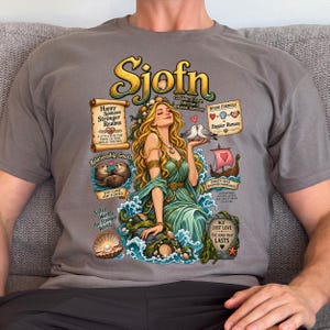 May include: A grey t-shirt featuring a graphic illustration of the goddess Sjöfn with long blonde hair, wearing a green dress, surrounded by sea-themed elements, two doves, otters, a ship, and text including "Happy Spouses Stronger Realms."
