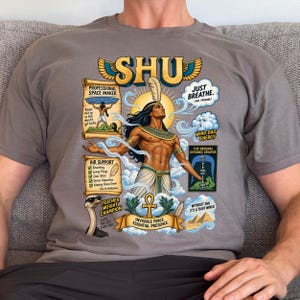 May include: A black t-shirt featuring a graphic of the Egyptian deity Shu. The design includes the text SHU, PROFESSIONAL SPACE MAKER, JUST BREATHE, AIR SUPPORT, FEATHER WEIGHT CHAMPION, and INVISIBLE FORCE. ESSENTIAL PRESENCE, with illustrations of clouds, an ankh, and pyramids.