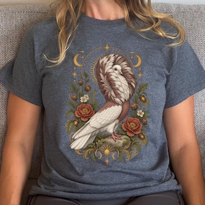 May include: A heathered dark blue t-shirt featuring a central graphic of a white and brown Jacobin pigeon perched on a rock. The bird is surrounded by orange flowers, green leaves, acorns, and gold celestial elements including moons and stars.